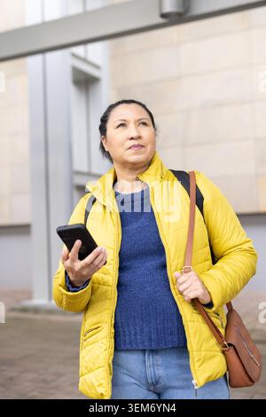 Standing woman in yellow coat, blue knit, holding smartphone, carrying brown bag by beige building Stock Photo