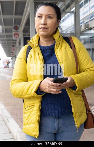 Mature woman standing at curbside, wearing yellow coat, checking smartphone while awaiting pickup Stock Photo