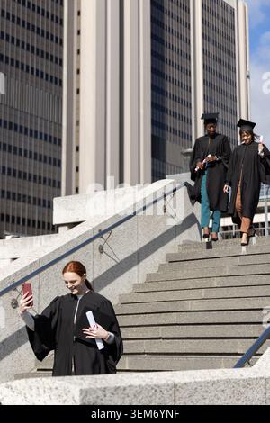 3 women grads walking steps by tower, in caps, with diplomas, taking phone selfie, copy space. Mortarboard, gown, scroll, trio, urban, skyscraper, sun Stock Photo
