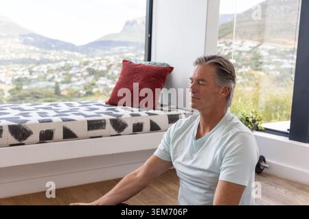 Senior man sitting meditating on wooden floor by large window with dumbbell, red pillow, plant Stock Photo