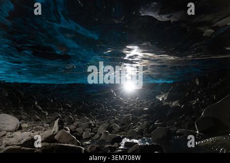 Interior of a glacial ice cave in Iceland with a bright star-burst light source illuminating the rocky ground and blue ice ceiling above, creating a d Stock Photo