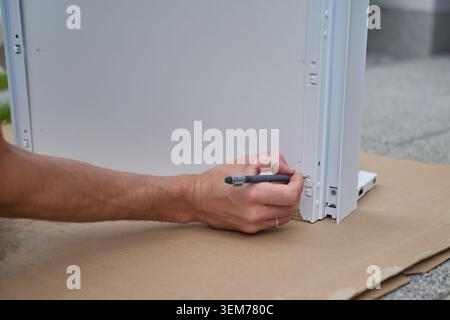 Man marks screw position with pencil on furniture panel during assembly. Measuring step before drilling and hardware installation Stock Photo