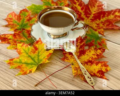 Coffee with cream in an antique cup with a silver gilded spoon on a saucer and an autumn maple leaf on an oak table. Close-up from the front. Stock Photo
