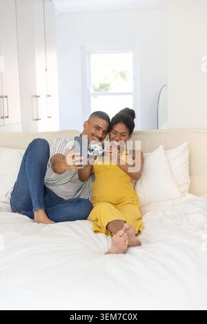 Expectant couple sitting on white bed in bedroom, woman in yellow holding ultrasound and smartphone Stock Photo