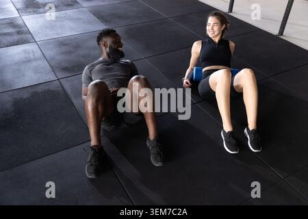 Diverse couple lying on rubber floor, woman wearing sneakers using blue foam roller by squat rack Stock Photo