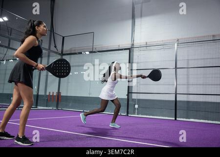 Incoming ball bouncing, player in white dress lunging to return shot, partner ready on purple court Stock Photo