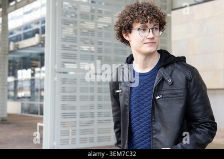 Teen male standing, looking sideways at plaza in leather jacket and backpack strap, copy space Stock Photo