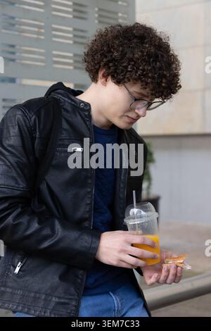Adolescent male standing at building facade holding orange drink and sandwich in leather jacket Stock Photo