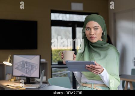 Adult woman in green hijab holding tablet, mug, looking in office at monitor showing 3D plan Stock Photo