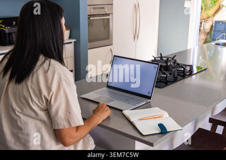 Mature woman in beige shirt sitting at kitchen island using laptop notebook and pencil, copy space Stock Photo