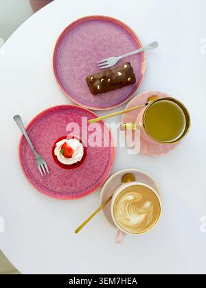 Vertical flat lay of coffee cup with latte art, green tea and mini cakes on pink plates Stock Photo