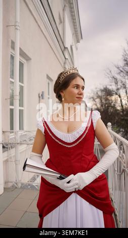 Young woman in beautiful red and white vintage ball gown, wearing a tiara and pearls, stands on a balcony. She looks around and smiles gracefully for Stock Photo