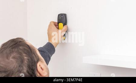 Mans hand using yellow cordless drill to fasten screw into white wall near black shelf bracket. Home improvement, additional securing, DIY reinforceme Stock Photo