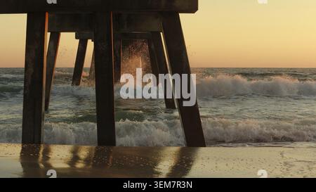 Waves are crashing against a wooden pier during sunset, creating a tranquil coastal scene with reflections on the wet sand and soft ocean hues Stock Photo