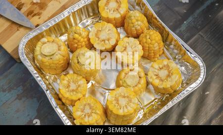 Fresh yellow corn cobs cut into halves are neatly arranged in a silver aluminum tray on a rustic wooden surface with a knife beside Stock Photo