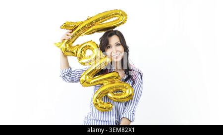 A young woman with long brown hair is joyfully holding a gold balloon shaped like the word love. She is smiling brightly and looking directly at the c Stock Photo