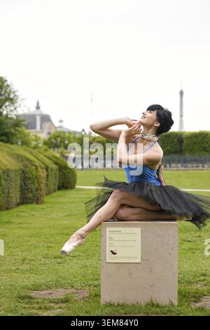 Graceful ballerina in black tutu and blue leotard performs expressive seated pose in a manicured garden with Eiffel Tower in background on a bright su Stock Photo