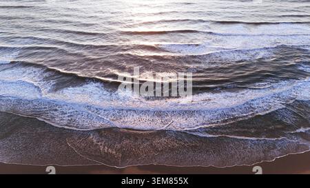 Top-down drone view captures textured ocean waves gently rolling onto the shoreline, glowing with warm sunset light and soft foam patterns Stock Photo