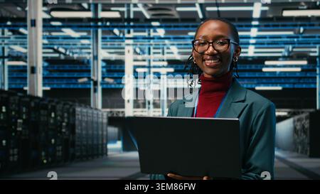 Portrait of happy data center system administrator doing resource optimization using laptop software. Smiling young employee in server hub using notebook to upgrade IT infrastructure, camera A Stock Photo