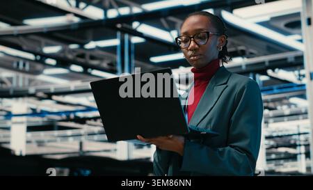 Managing databases, laptop and female technician walking in data center with augmented reality storage rigs. Cloud computing, VFX overlay and African american woman working in server farm, camera B Stock Photo