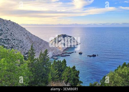 Steep rocky cliff on the left descends to a small, tree-covered peninsula surrounded by clear blue waters. The calm sea reflects pastel hues from the Stock Photo