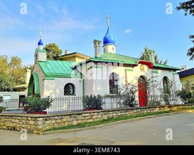 A small Russian-style church with distinctive architectural features, including a blue onion dome and green roofs, is surrounded by a modest iron fenc Stock Photo