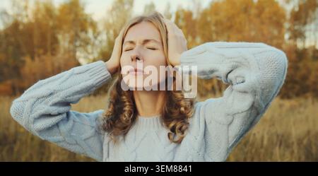 Upset exhausted young woman with headache, pain tension in the head, suffers from migraine standing outdoors in autumn park Stock Photo