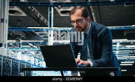 Data center admin typing on laptop keyboard, doing resource optimization using software. Server farm IT specialist using notebook program to upgrade hardware supercomputers, camera A Stock Photo