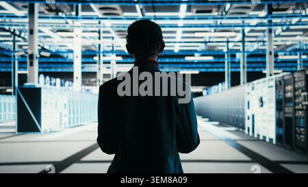 Software developer walks in data center, using tablet and AR technology to debug system errors. Server farm IT professional runs diagnostics on blue wireframe mesh equipment supercomputers, camera A Stock Photo