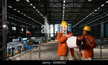 Diverse team of engineers studying blueprints and layout plans in machinery hall, man and woman checking instructions and circuits for heavy equipment to ensure production workflow. Stock Photo