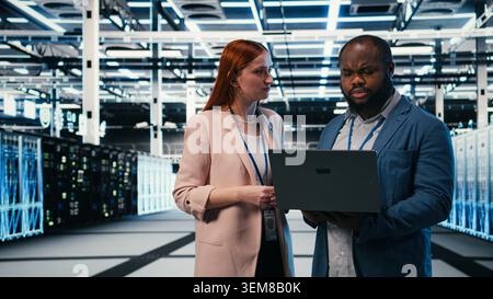 Optimizing electronics, laptop and manager teaching intern in server farm during induction. Monitoring system performance, notebook and team leader with trainee in data center, camera A Stock Photo