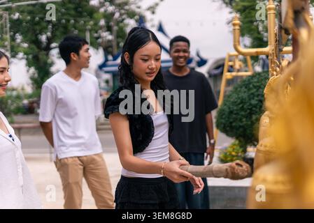 Young asian woman ringing golden temple bells, experiencing thai spirituality with friends Stock Photo