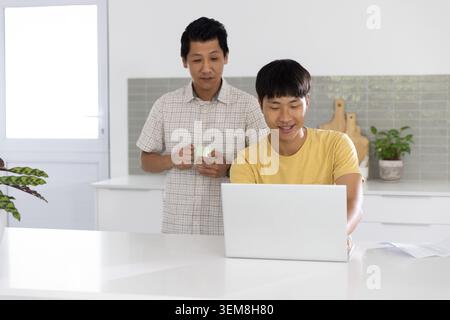 Asian father holding light green mug while son smiling and using silver laptop at kitchen island Stock Photo