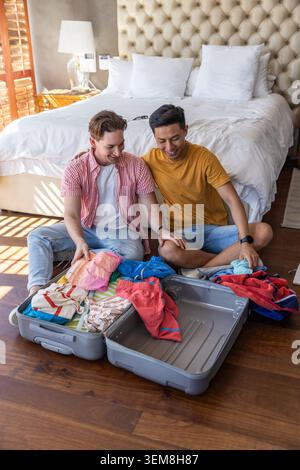 Diverse male partners sitting on hardwood floor unpacking gray hard-shell suitcase in bedroom. Couple, packing, luggage, sunlight, hotelroom, cozy, ca Stock Photo