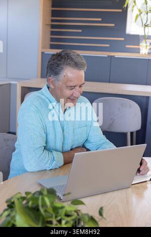 Man in turquoise button-up shirt working on silver laptop while writing on notepad at kitchen table Stock Photo