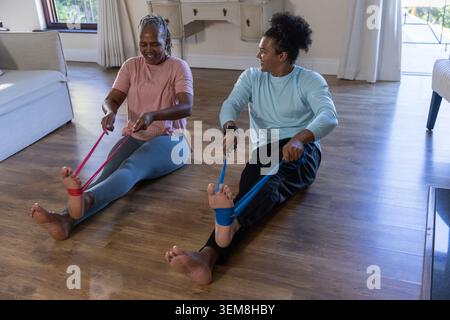 Man and senior African American woman stretching legs using resistance bands on living room floor Stock Photo