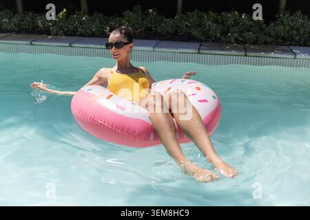 Female reclining on pink sprinkle donut float in pool, wearing yellow one-piece and dark shades Stock Photo
