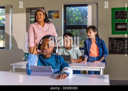 Diverse classroom of elementary students and teacher sitting at desks using tablets and headphones Stock Photo