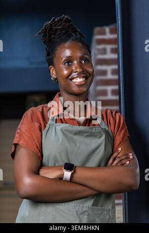 Adult African American woman standing with arms crossed in cafe wearing green apron and smartwatch. Barista, brick, workspace, smiling, warm, casual, Stock Photo