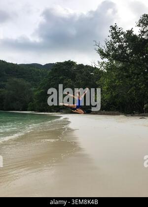 Beautiful young woman performing an elegant mid-air leap on a pristine tropical beach, capturing the grace of a dancer and the athleticism of gymnast. Stock Photo