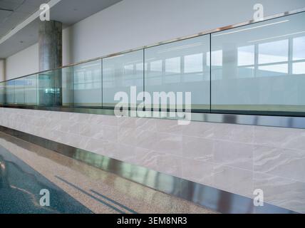 Glass barrier panels above the tiled stone wall base stretch along the empty passage corridor inside the building hall to guide pedestrian traffic. Stock Photo
