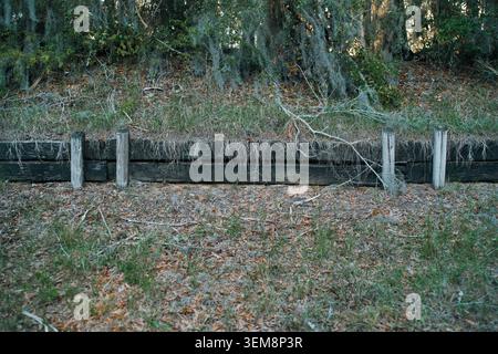 Old dark wooden retaining wall constructed from heavy timber logs holding back an earthen bank covered with dry grass and fallen forest leaves. Stock Photo