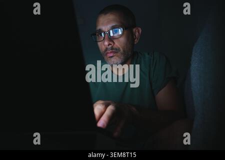 Man focusing on his laptop screen, illuminated by its reflection on his glasses in a dark setting Stock Photo