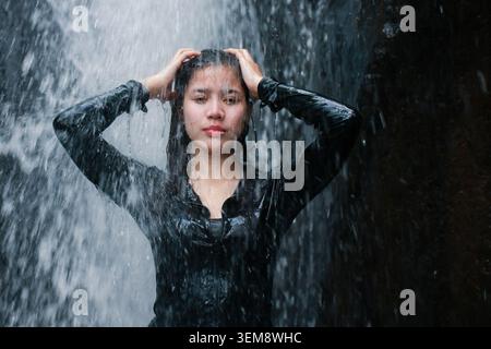 Young woman standing under waterfall touching hair relaxing outdoors enjoying nature tropical environment feeling calm peaceful during heavy rain wet Stock Photo