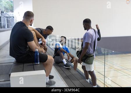 Diverse male teammates chatting on benches by glass rail over gym court, duffel and water bottle Stock Photo