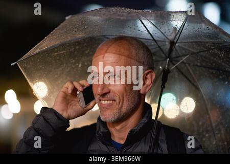 Outdoor, umbrella and man with phone call at night, digital communication and commute for networking. Smile, rain and mature person with mobile for Stock Photo