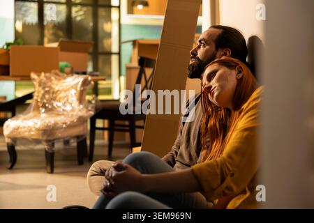 Homeowners couple, sitting on floor, proud of new house, buying their first home, and taking a break from unpacking things. Young people tired but content with their new beginning together. Stock Photo