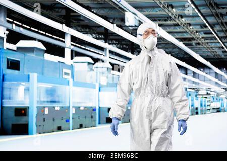 Engineer man walking in microchip manufacturing plant laboratory, wearing protective gear. Integrated circuits industrial factory professional in cleanroom checking automated machinery yield output Stock Photo