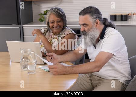 Senior couple sitting at round wooden table in kitchen, reviewing laptop and scattered documents Stock Photo