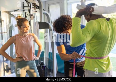 Diverse trio doing waist check in gym, trainer wrapping pink tape, client holding white towel Stock Photo